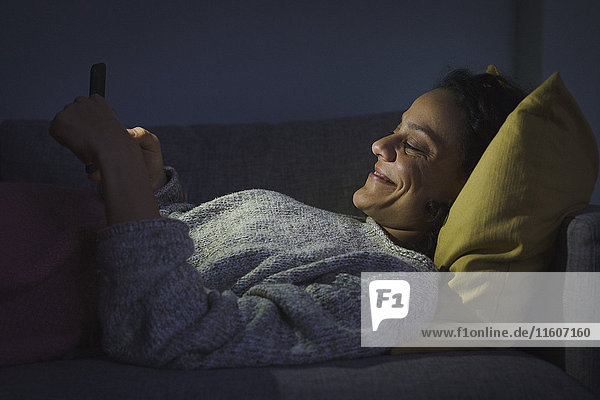 Seitenansicht der glücklichen Frau mit dem Smartphone auf dem Sofa im dunklen Wohnzimmer