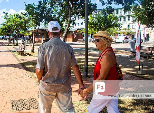 Older mature retired caucasian couple holding hands in the streets of Fort de France on a vacation. He wears a baseball hat and she a straw hat.