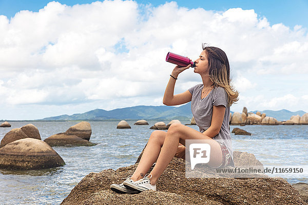 Teenager-Mädchen sitzt auf Felsen und trinkt aus Wasserflasche