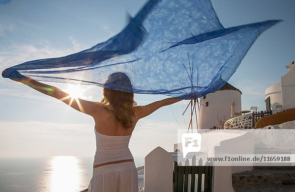 Frau hebt die Arme und hält einen Kaftan in der Hand und blickt weg auf das Meer  Santorin  Kykladen  Griechenland