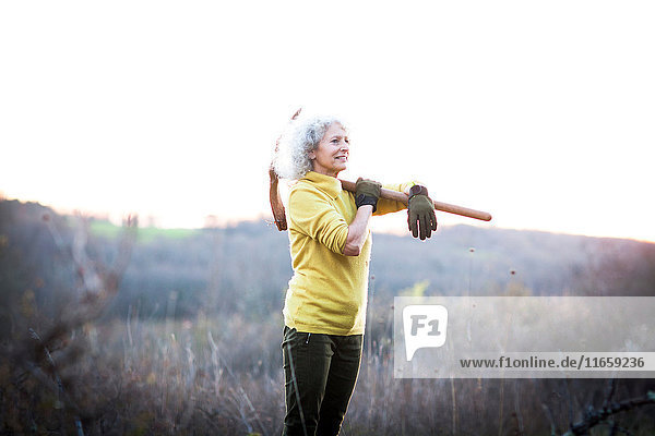 Reife Frau trägt in der Landschaft eine Spitzhacke über der Schulter