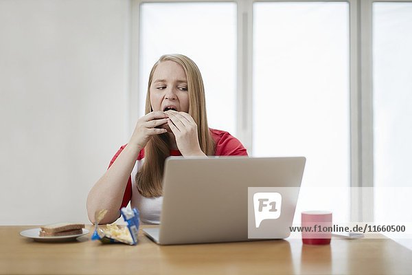 Junge Frau mit Laptop  die ein Sandwich isst.