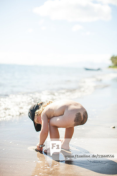 Junge steht am Strand  bückt sich  gräbt im Sand  Sand auf nacktem Gesäß