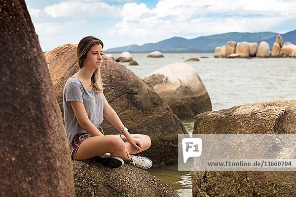 Teenager-Mädchen sitzt im Schneidersitz auf Felsen am Meer und schaut weg