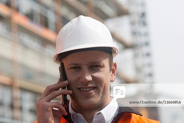 Porträt eines jungen männlichen Bauingenieurs  der auf einer Baustelle mit einem Smartphone telefoniert