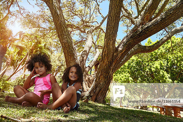 Portrait of two young sisters  sitting beside tree