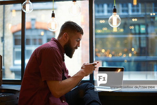 Junger männlicher Hipster schaut auf Smartphone im Cafe Fenstersitz