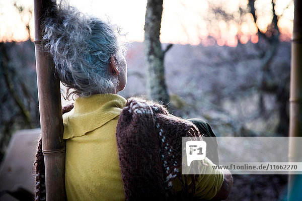 Rückansicht einer reifen Frau,  die auf einer Waldveranda sitzt und den Sonnenuntergang betrachtet