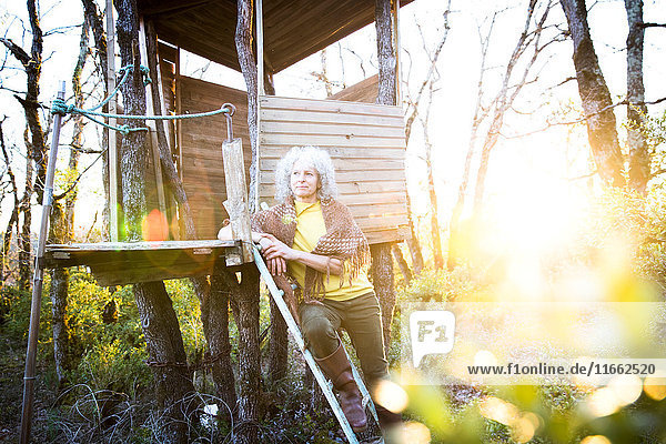 Reife Frau lehnt bei Sonnenuntergang an Baumhausleiter im Wald