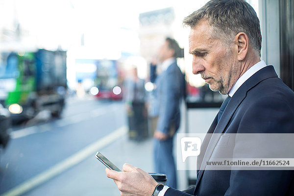 Geschäftsmann wartet an der Bushaltestelle mit Smartphone  London  UK