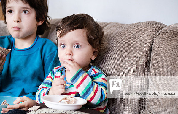 Kleiner Junge und großer Bruder sitzen auf dem Sofa,  sehen fern und essen einen Snack