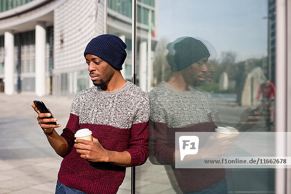 Junger Mann  im Freien  am Fenster lehnend  mit Smartphone  mit Einweg-Kaffeetasse