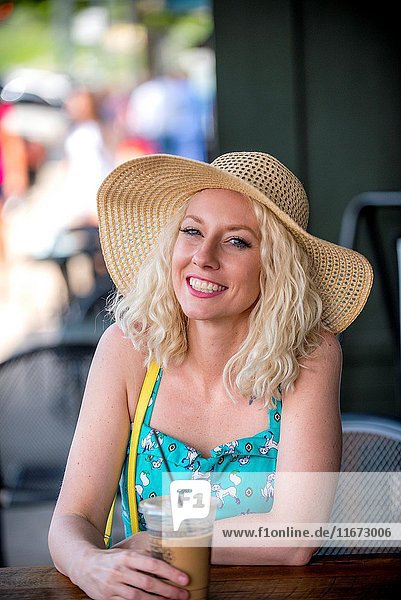 A pretty 30 year old blond woman sitting at an outdoor cafe with a cold drink smiling at the camera.