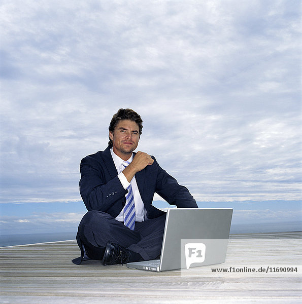 Ein Mann in Anzug und Krawatte sitzt auf einem Deck am Meer und arbeitet an einem Laptop.