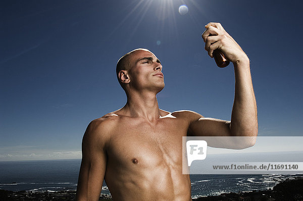 Ein Mann ohne Hemd steht vor dem Ozean und hält eine Flasche Sonnencreme in der Luft.