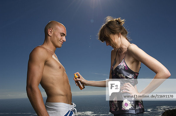 Mann und Frau ohne Hemd stehen vor dem Meer  Frau trägt Sonnencreme auf den Bauch des Mannes auf.