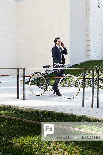 Mittlerer erwachsener Geschäftsmann  der ein Fahrrad hält und ein Mobiltelefon benutzt