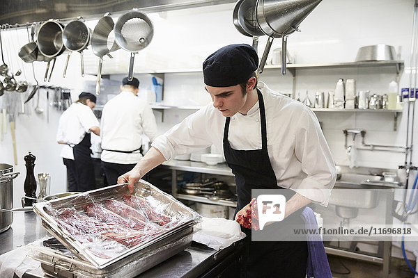 Männlicher Küchenchef  der in der Großküche ein mit Plastik umwickeltes Tablett mit Fleisch untersucht.