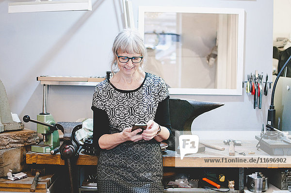 Smiling senior woman using smart phone in jewelry workshop