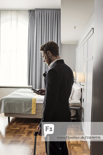 Side view of businessman using mobile phone standing with luggage in hotel room