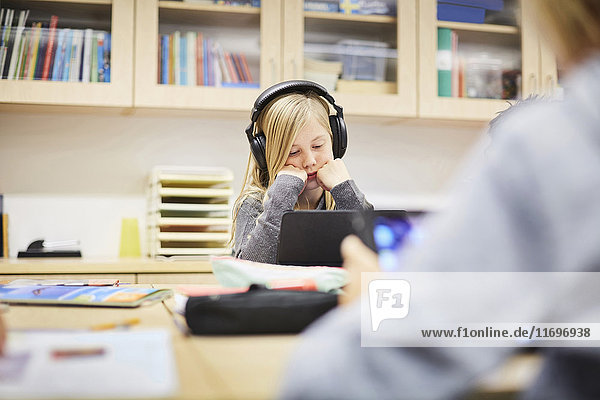 Girl wearing headphones while looking at digital tablet at desk in classroom