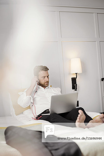 Ein reifer Geschäftsmann  der im Hotelzimmer auf dem Bett sitzt und den Kopfhörer am Laptop einstellt.
