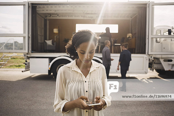 Geschäftsfrau mit Smartphone mit Kollegen und tragbarem Bürowagen im Hintergrund
