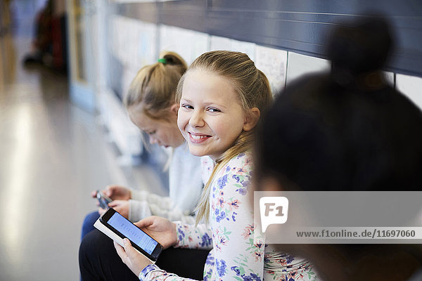 Lächelndes Mädchen hält Smartphone  während es einen Freund im Schulflur ansieht.