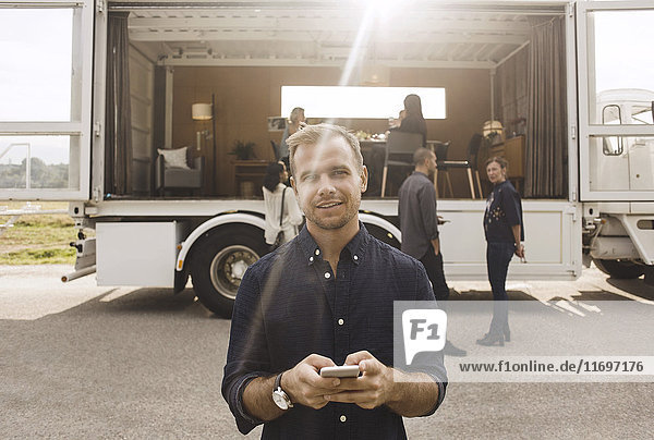 Porträt eines Geschäftsmannes  der sein Handy mit Kollegen und einem tragbaren Bürowagen im Hintergrund hält.