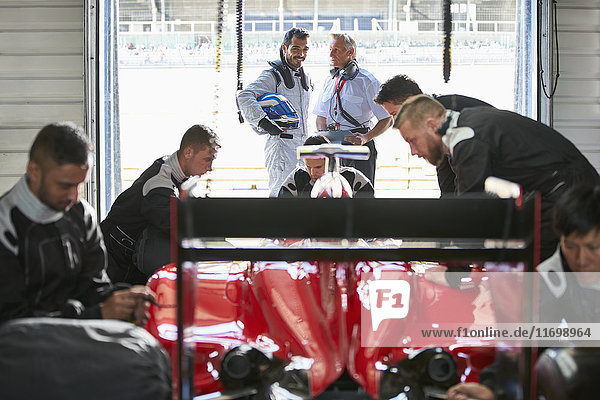 Formula one driver and manager talking behind pit crew working on race car in repair garage