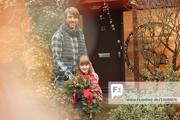 Portrait lächelnder Vater und Tochter mit Weihnachtskranz vor dem Haus