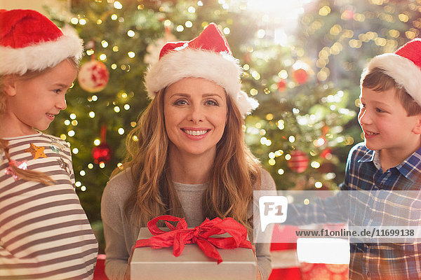 Porträt einer lächelnden Mutter mit Weihnachtsmannmütze  die ein Weihnachtsgeschenk von Tochter und Sohn erhält