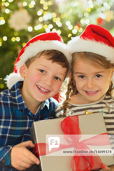 Porträt lächelnder Bruder und Schwester mit Weihnachtsmannmützen  die ein Weihnachtsgeschenk halten