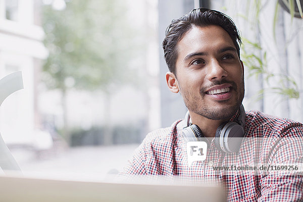 Lächelnder junger Mann mit Kopfhörern am Laptop im Caféfenster
