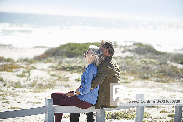 Affectionate senior couple sitting on sunny beach fence