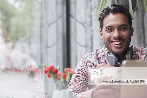 Porträt eines lächelnden jungen Mannes mit Kopfhörern  der Kaffee am Laptop im Caféfenster trinkt