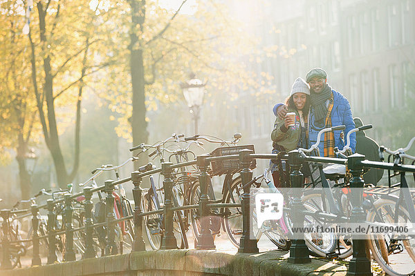 Portrait smiling young couple drinking coffee along bicycles on sunny urban autumn bridge  Amsterdam