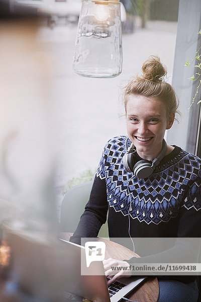 Porträt einer lächelnden jungen Frau,  die in einem Caféfenster einen Laptop benutzt