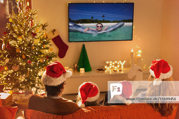 Familie mit Weihnachtsmannmützen sieht sich im Weihnachtswohnzimmer ein Sommerferienvideo im Fernsehen an