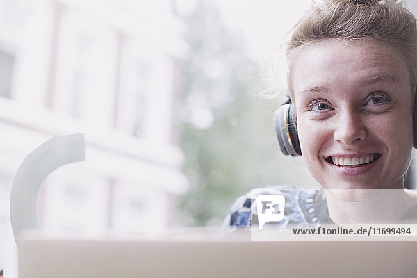 Porträt einer lächelnden jungen Frau mit Kopfhörern am Laptop in einem Caféfenster