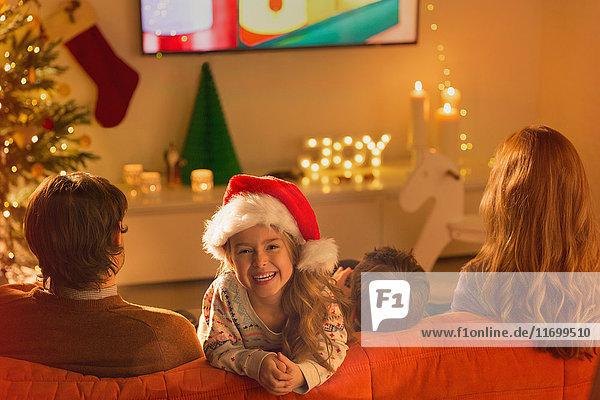 Porträt eines lächelnden Mädchens mit Weihnachtsmannmütze  das mit seinen Eltern im Weihnachtswohnzimmer fernsieht