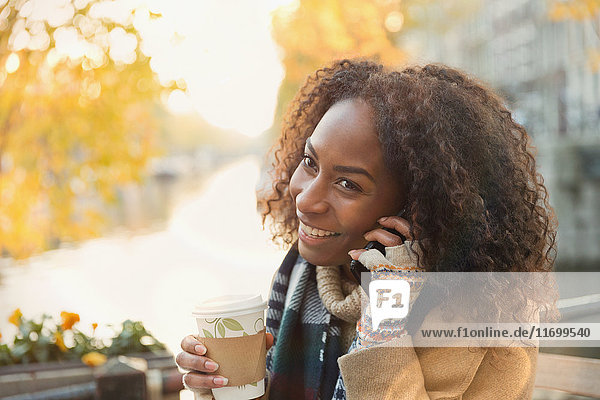 Smiling young woman drinking coffee and talking on cell phone at autumn sidewalk cafe