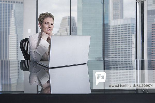 Lächelnde Geschäftsfrau am Schreibtisch im Stadtbüro mit Blick auf den Laptop