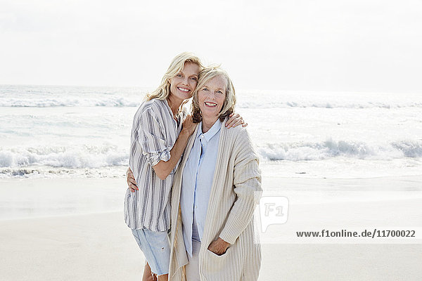 Seniorin und ihre erwachsene Tochter stehen am Strand und umarmen sich.