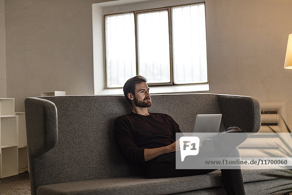 Young man sitting with laptop on couch looking at distance