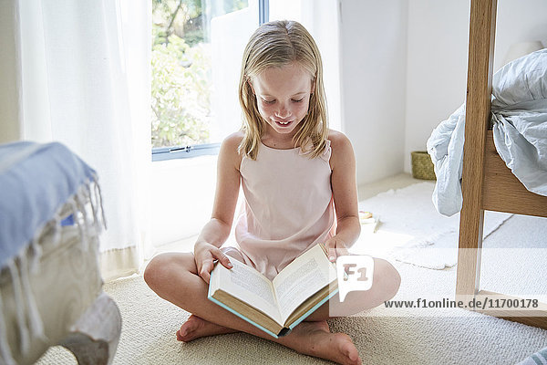 Kleines Mädchen,  das zu Hause auf dem Boden sitzt und ein Buch liest.
