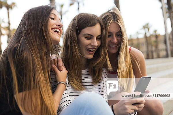 Drei glückliche junge Frauen sitzen auf der Treppe und haben Spaß mit dem Handy.