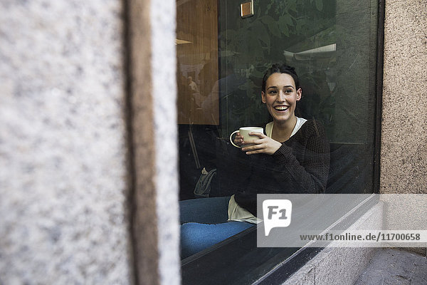 Porträt einer lächelnden Frau mit einer Tasse Kaffee hinter einer Fensterscheibe in einem Coffee-Shop.