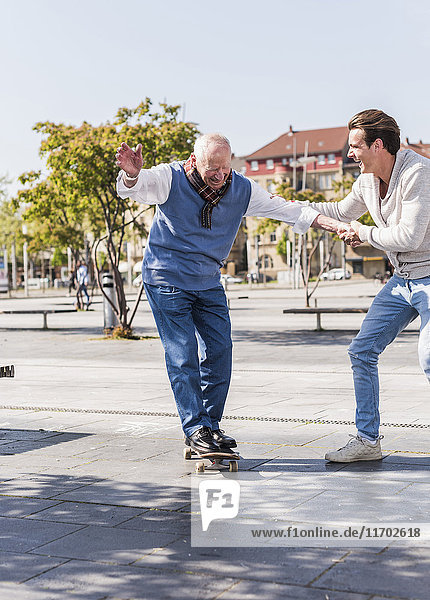 Erwachsener Enkel  der dem älteren Mann auf dem Skateboard assistiert.