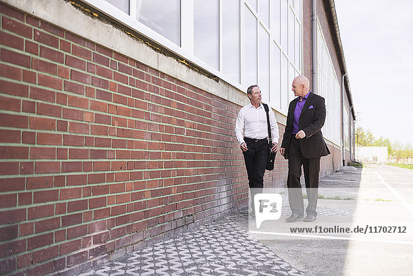 Two men walking along factory building talking